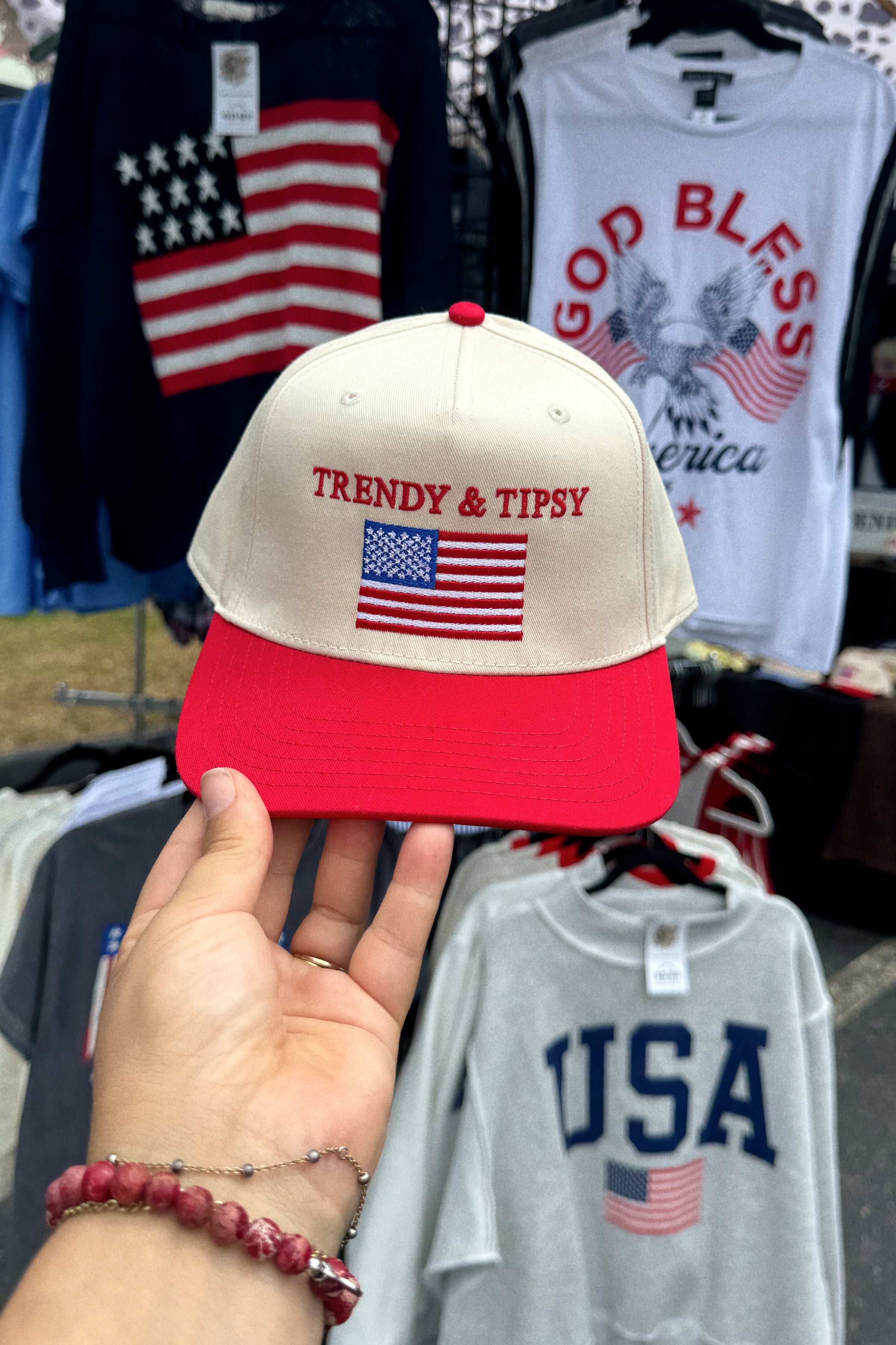 Beige and red cap with 'TRENDY & TIPSY' and American flag design held in front of clothing items.