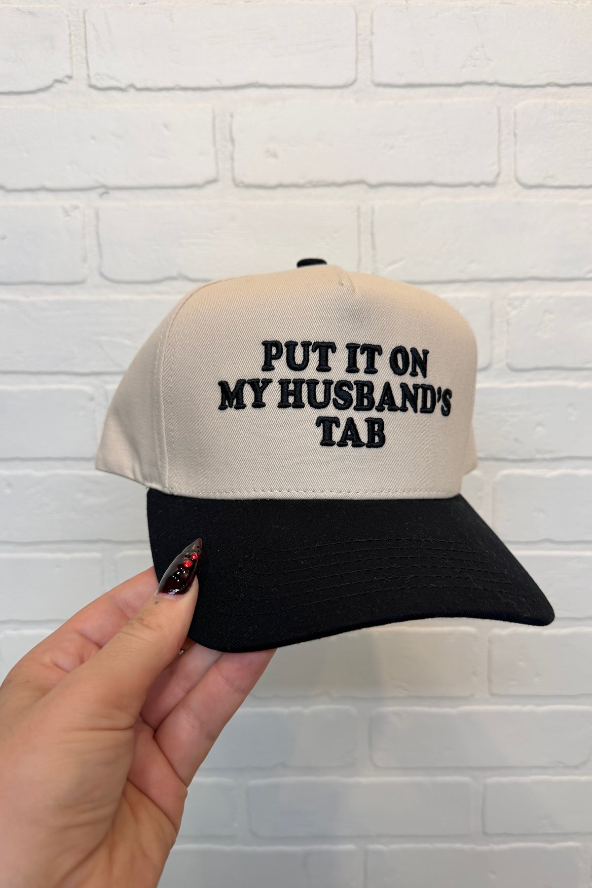 A hand holding a beige and black trucker-style cap with the embroidered text 'PUT IT ON MY HUSBAND'S TAB' against a white brick wall background.