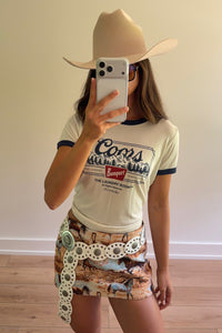 A woman takes a mirror selfie wearing a beige cowboy hat, a cream ringer T-shirt with dark blue trim and a graphic print, and a brown camouflage mini skirt with a decorative white belt featuring silver-toned conchos.