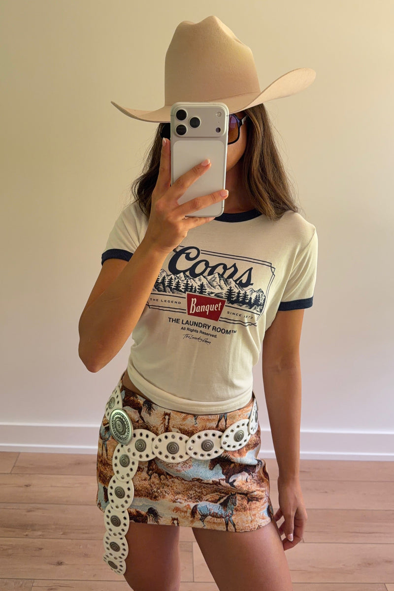 A woman takes a mirror selfie wearing a beige cowboy hat, a cream ringer T-shirt with dark blue trim and a graphic print, and a brown camouflage mini skirt with a decorative white belt featuring silver-toned conchos.