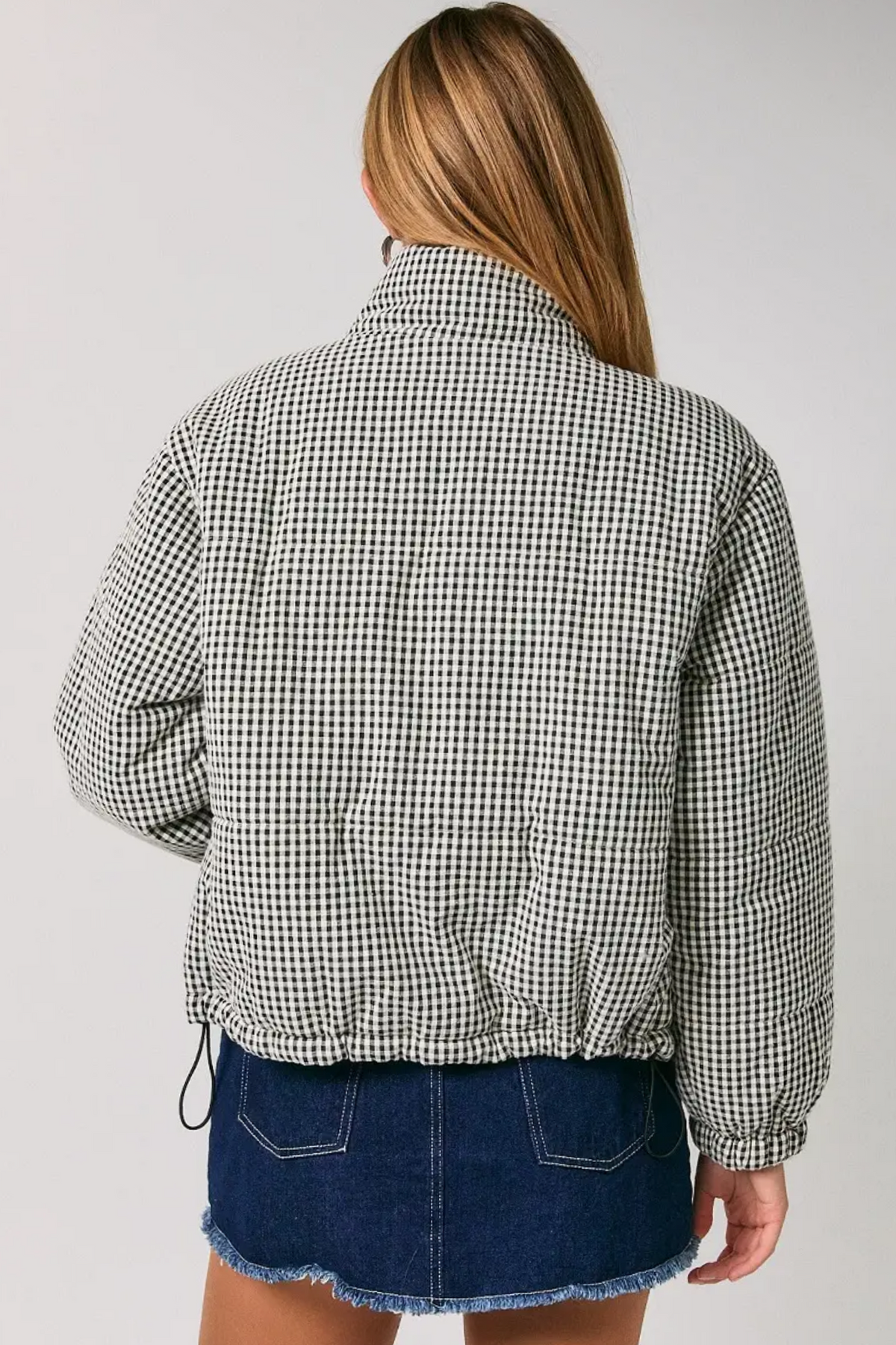 A woman shown from the back wearing a black and white gingham patterned cropped jacket and a blue denim skirt against a plain light gray studio background.