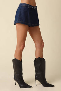 A model's lower body wearing a dark blue denim mini skirt with a matching belt and black pointed-toe cowboy boots, set against a solid beige background.