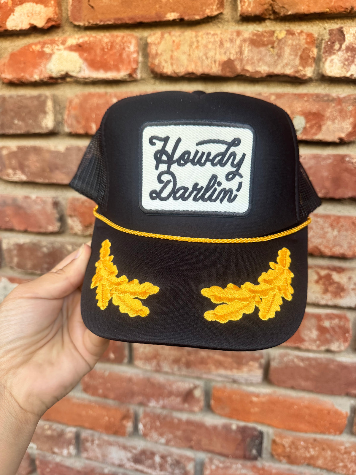 A hand holds a black trucker hat in front of a red brick wall. The hat features a white rectangular patch with the words 'Howdy Darlin'' in black script, a yellow embroidered stripe across the brim, and two yellow embroidered leaf designs on the sides.