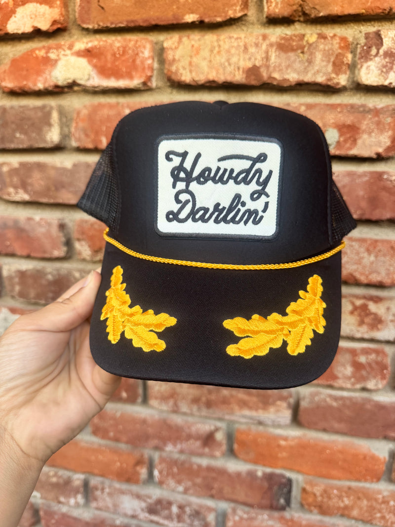 A hand holds a black trucker hat in front of a red brick wall. The hat features a white rectangular patch with the words 'Howdy Darlin'' in black script, a yellow embroidered stripe across the brim, and two yellow embroidered leaf designs on the sides.