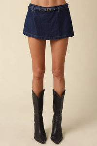 A model's lower body wearing a dark blue denim mini skirt with a matching belt and black pointed-toe cowboy boots, set against a solid beige background.