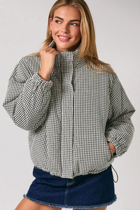 A woman wearing a black and white gingham patterned puffer jacket with a high collar, paired with a dark denim mini skirt, against a light gray studio background.