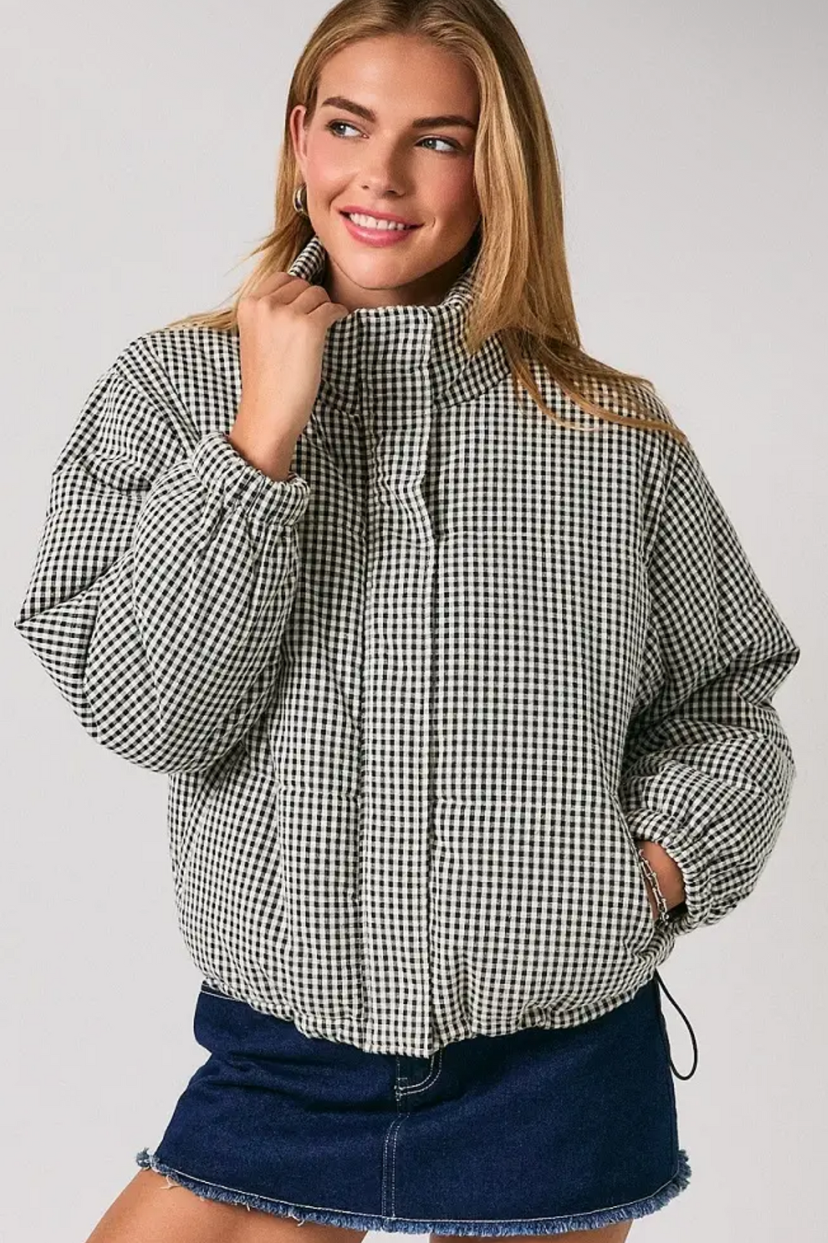 A woman wearing a black and white gingham patterned puffer jacket with a high collar, paired with a dark denim mini skirt, against a light gray studio background.