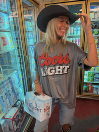 A woman wearing a black cowboy hat and a gray Coors Light graphic t-shirt stands in front of a Coors refrigerator, holding a Coors Light cardboard cooler bag.
