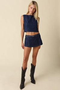A model's lower body wearing a dark blue denim mini skirt with a matching belt and black pointed-toe cowboy boots, set against a solid beige background.