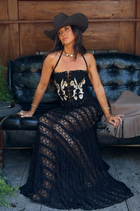 A woman wearing a black lace maxi dress with a corset-style bodice, a wide-brimmed black cowboy hat, and gold jewelry, sitting on a black leather tufted couch against a wooden wall.