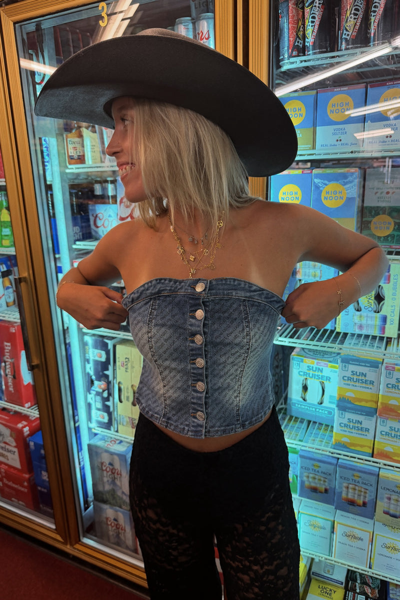 A woman wearing a blue denim strapless tube top with front buttons, black lace pants, and a large black cowboy hat, standing in front of a retail refrigerator.