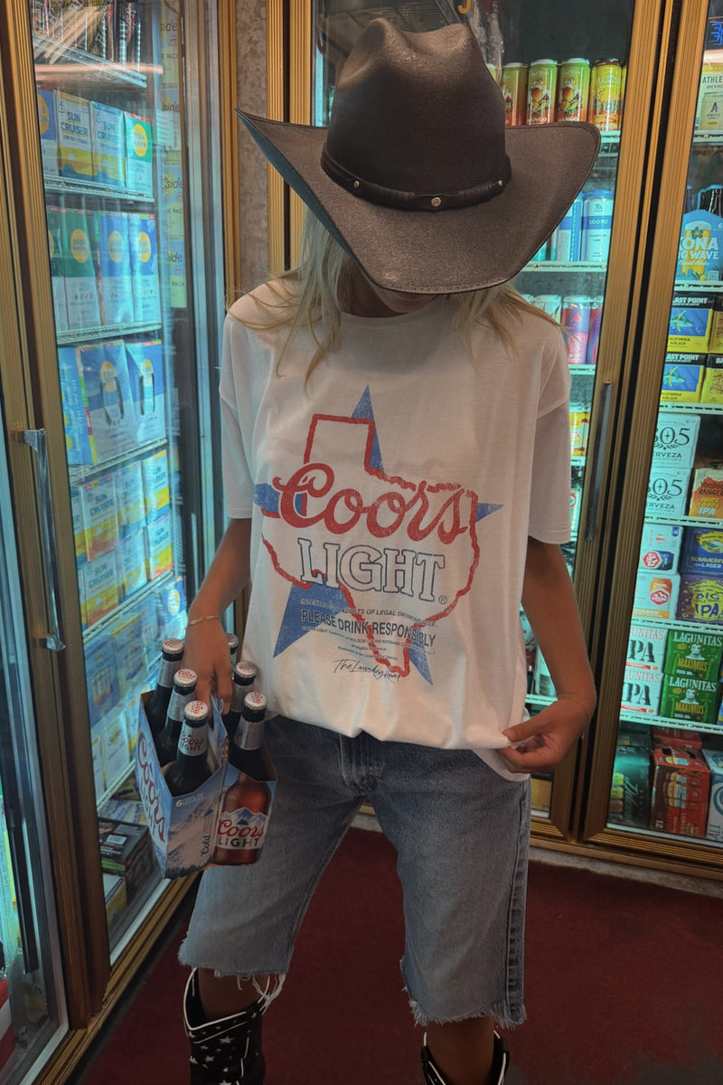 A person standing in a retail store in front of beverage glass-door refrigerators, wearing an oversized white Coors Light graphic T-shirt, a brown cowboy hat, light-wash denim shorts, and black boots while holding a six-pack of beer.