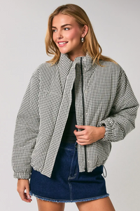 A woman modeling a black and white houndstooth patterned puffer jacket over a black top and a blue denim mini skirt, photographed against a solid light gray studio background.