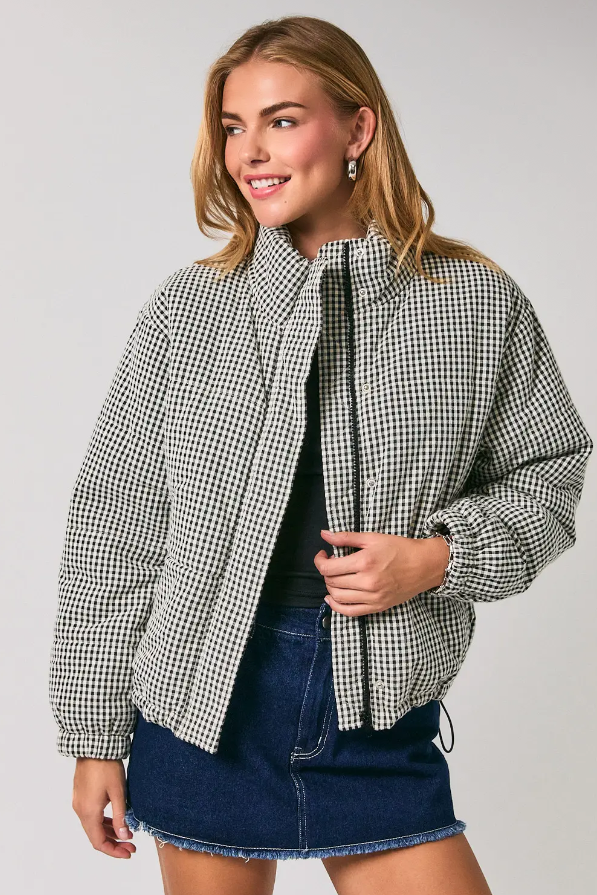 A woman modeling a black and white houndstooth patterned puffer jacket over a black top and a blue denim mini skirt, photographed against a solid light gray studio background.