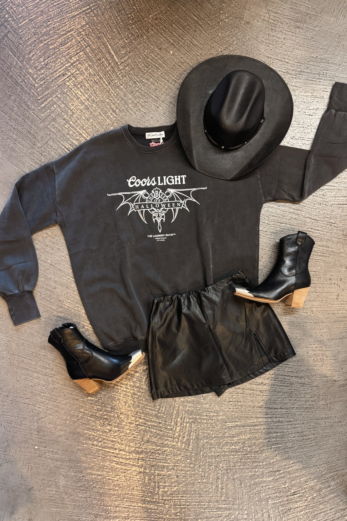 A flat-lay outfit arrangement on a textured gray floor featuring a black Coors Light sweatshirt with a white graphic, black leather-look shorts, a black cowboy hat, and black ankle boots with light wood heels.