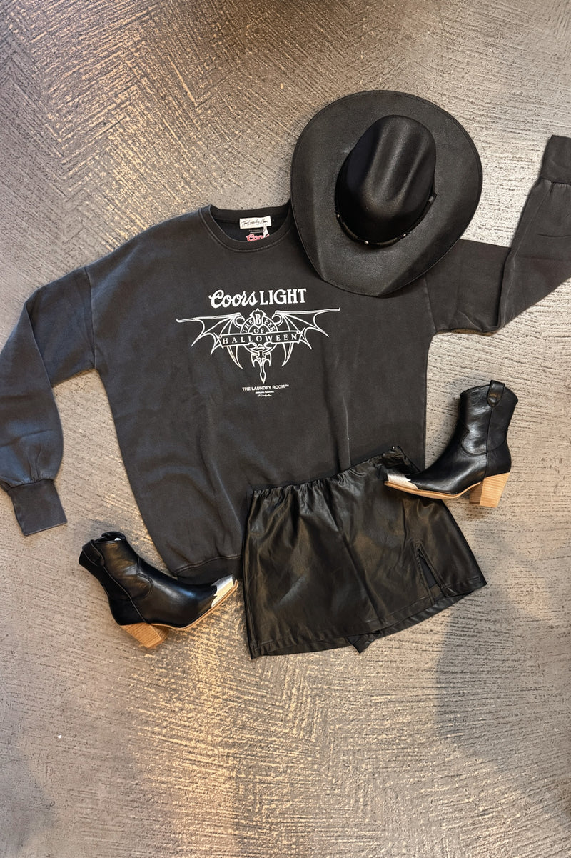 A flat-lay outfit arrangement on a textured gray floor featuring a black Coors Light sweatshirt with a white graphic, black leather-look shorts, a black cowboy hat, and black ankle boots with light wood heels.