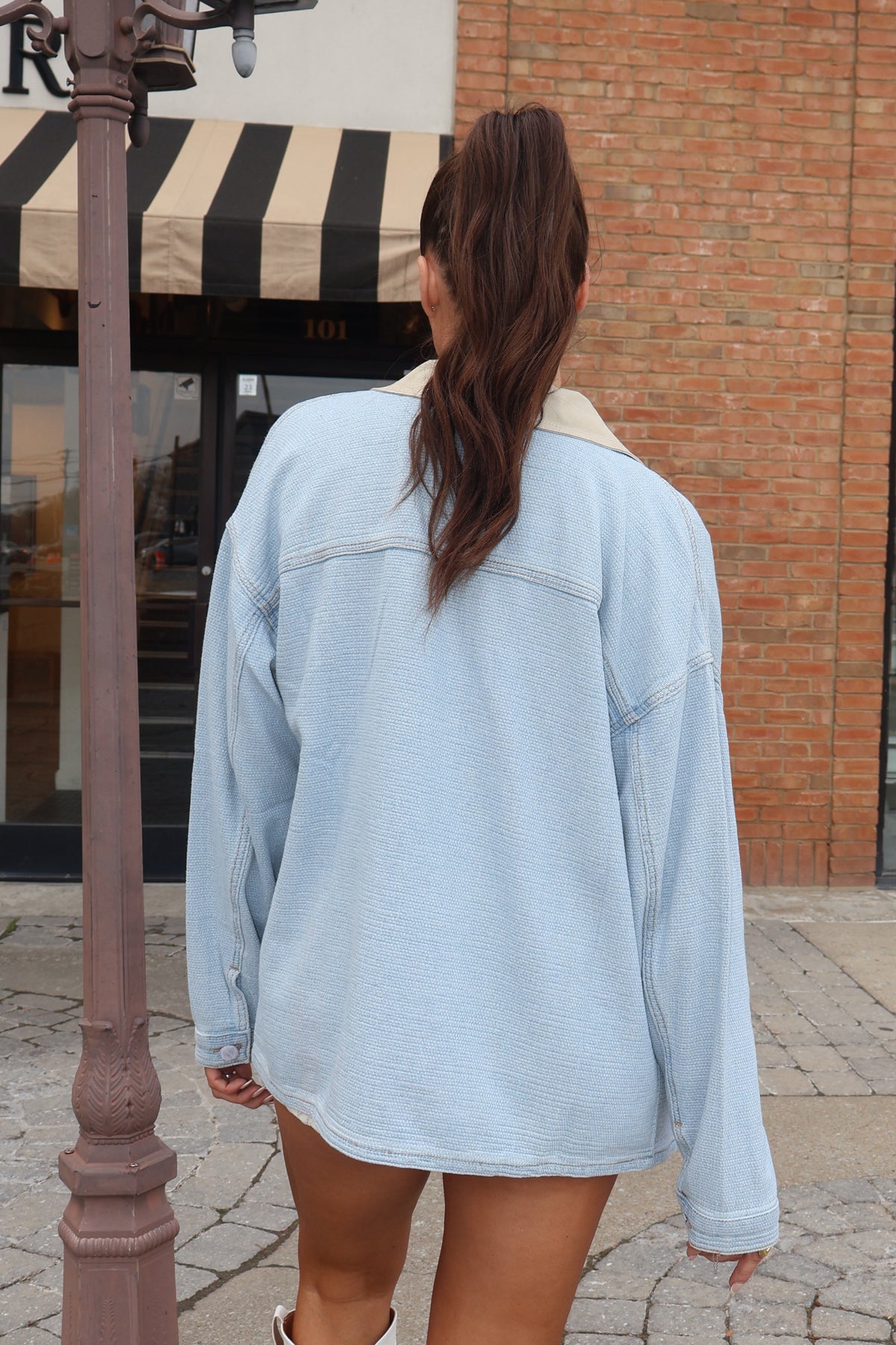 Person wearing a light blue denim jacket with a brick building and striped awning in the background