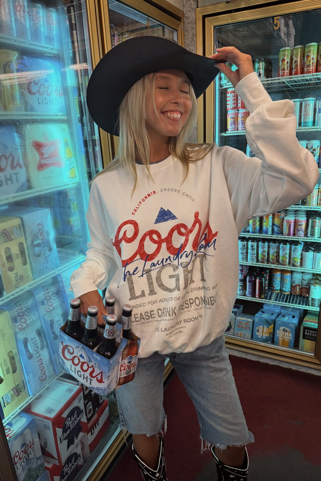 A woman wearing a black cowboy hat and a white Coors Light sweatshirt stands inside a retail store near beverage coolers, holding a six-pack of Coors Light beer.