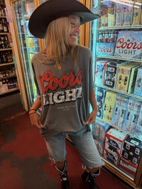 A woman wearing a Coors Light graphic t-shirt, denim shorts, and a black cowboy hat standing in front of a Coors Light refrigerator in a retail store.