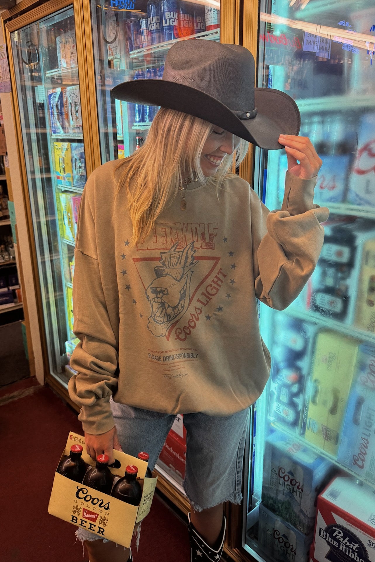 A woman wearing a beige graphic sweatshirt, denim shorts, a wide-brimmed dark hat, and black boots stands in front of a refrigerator in a retail store, holding a Coors beer case.