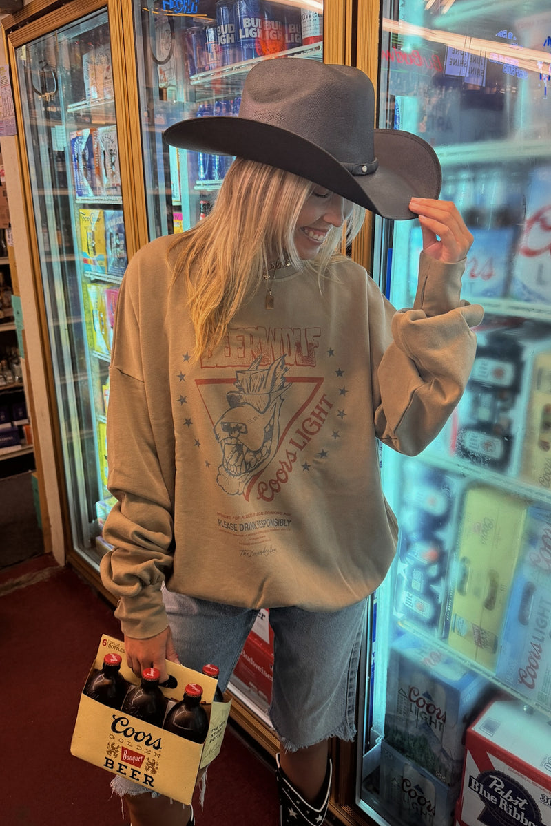 A woman wearing a beige graphic sweatshirt, denim shorts, a wide-brimmed dark hat, and black boots stands in front of a refrigerator in a retail store, holding a Coors beer case.