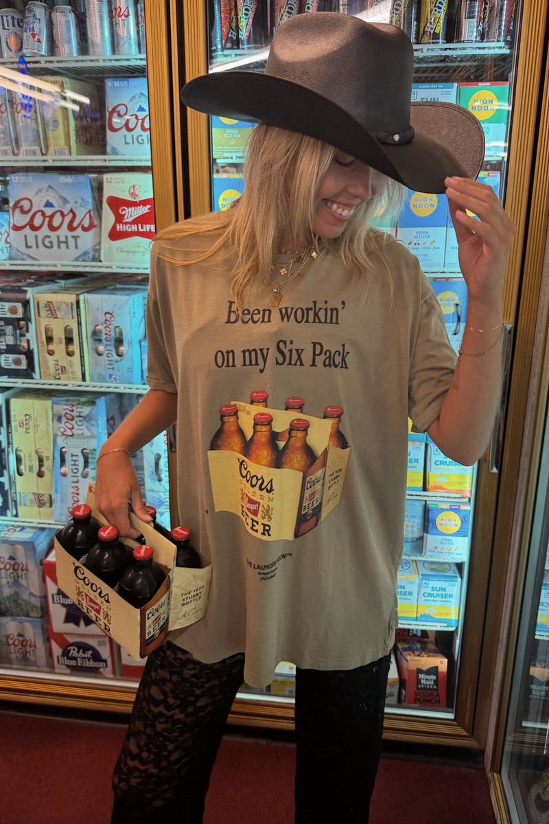 A woman wearing a brown cowboy hat and a beige graphic t-shirt that reads 'Been workin' on my Six Pack' with a beer carton print, holding a six-pack of Coors Lager in front of a refrigerator filled with beer bottles.