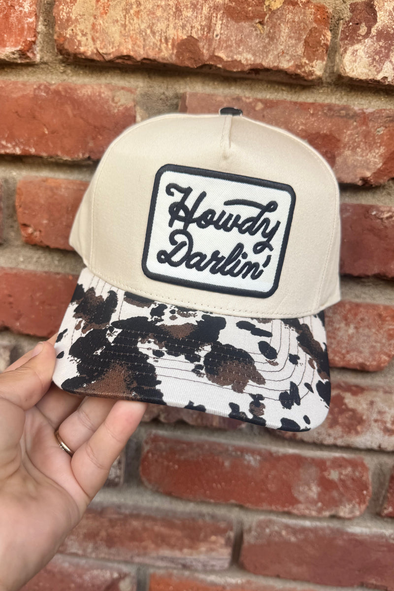 A beige snapback cap with a black and white cow-print brim, held by a hand against a red brick wall. The front of the cap features a square patch with the text 'Howdy Darlin''.