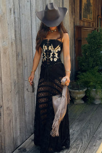 A woman wearing a black strapless maxi dress with a corset-style bodice, lace skirt, and fringe details, styled with a wide-brimmed gray hat, gold jewelry, and a light beige fringed clutch, standing on a rustic wooden porch.