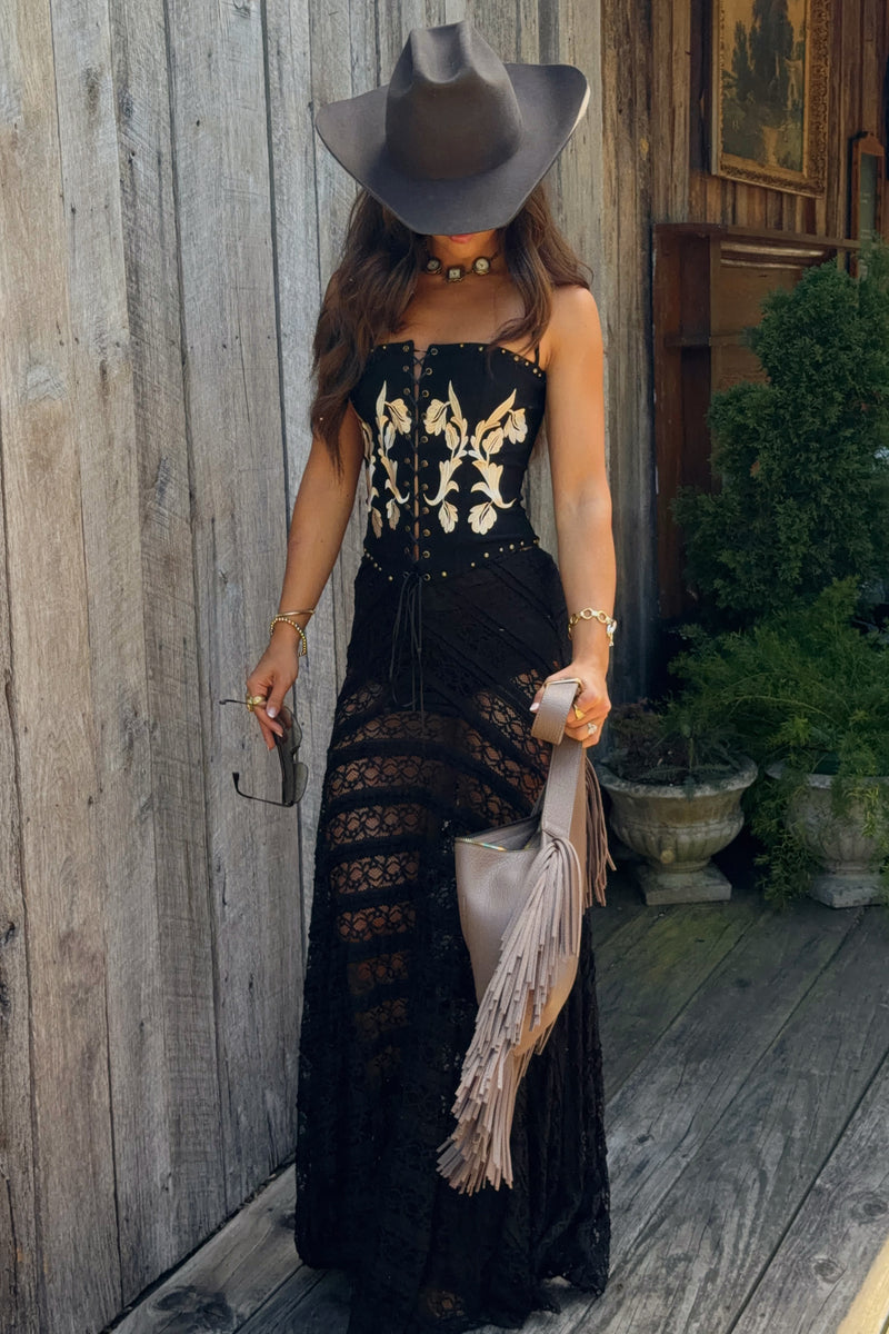 A woman wearing a black strapless maxi dress with a corset-style bodice, lace skirt, and fringe details, styled with a wide-brimmed gray hat, gold jewelry, and a light beige fringed clutch, standing on a rustic wooden porch.