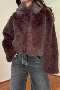 A person wearing a cropped, dark brown faux fur jacket with a matching collar, paired with gray jeans, standing indoors against a plain wall.