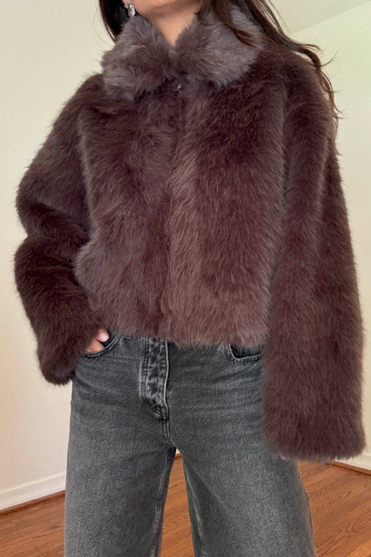 A person wearing a cropped, dark brown faux fur jacket with a matching collar, paired with gray jeans, standing indoors against a plain wall.