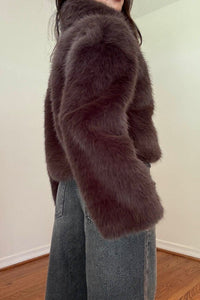 A person wearing a dark brown faux fur cropped jacket and wide-leg gray jeans, standing in an indoor room with a light-colored wall and wooden floor.