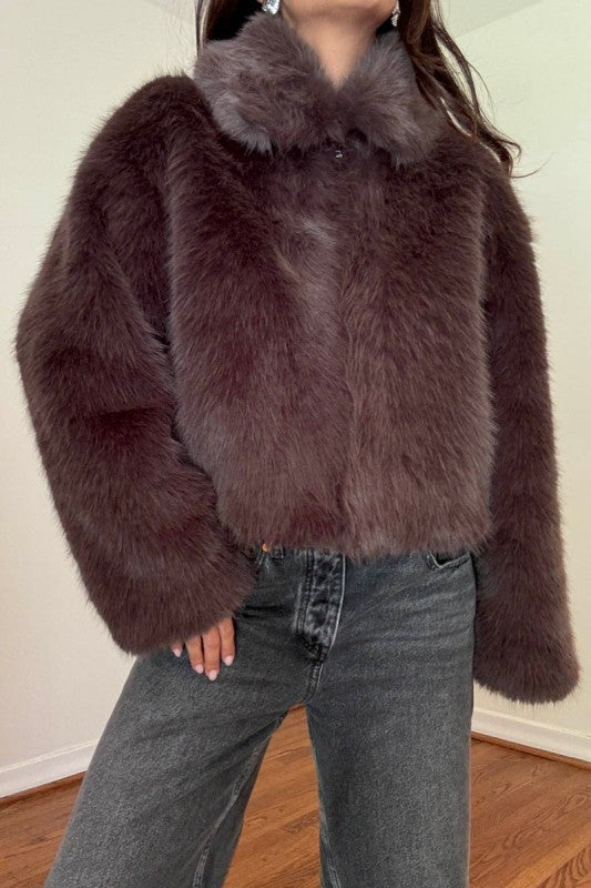 A person wearing a dark brown shaggy faux fur cropped jacket with a wide collar, paired with gray jeans, standing indoors against a plain wall.