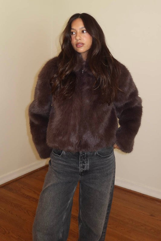 A woman wearing a dark brown faux fur cropped jacket and dark gray jeans, standing in a room with a beige wall and wooden floor.