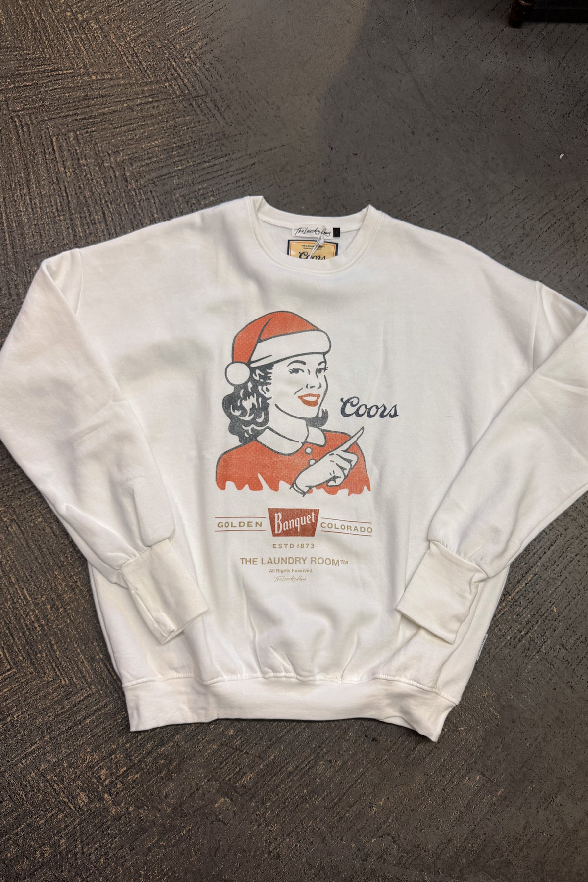 A white crewneck sweatshirt featuring a vintage-style Coors advertisement graphic with a woman in a Santa hat, laid flat on a dark textured carpet.