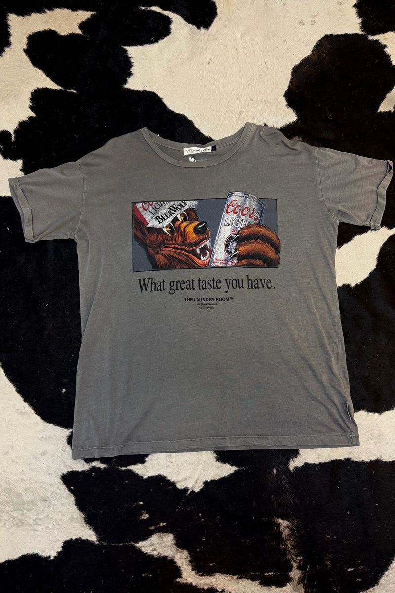 A gray short-sleeve graphic t-shirt laid flat on a black and white cowhide rug. The graphic features a bear holding a Coors can with the text 'What great taste you have.