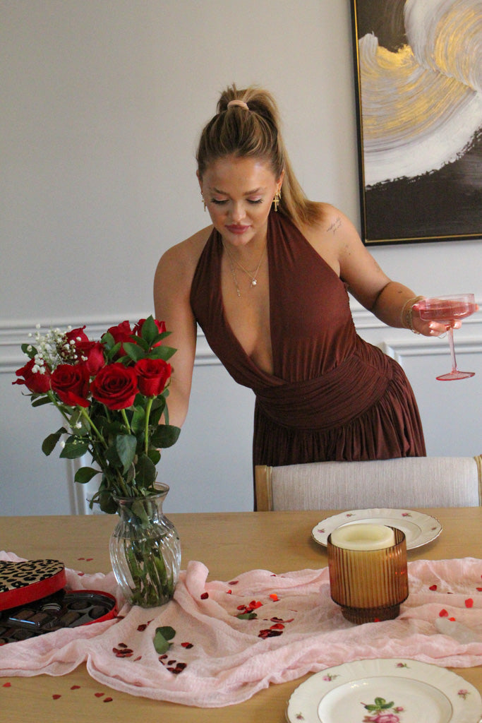 Woman in a brown dress with a glass of pink wine, surrounded by red roses and decorative items on a table.