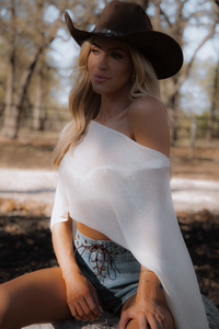 Woman wearing a white off-shoulder top and denim shorts with a brown cowboy hat, sitting outdoors.
