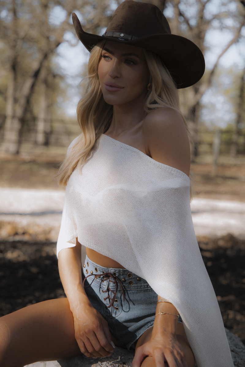 Woman wearing a white off-shoulder top and denim shorts with a brown cowboy hat, sitting outdoors.
