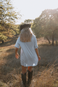 Person wearing a white dress and black boots walking through a field with trees in the background.