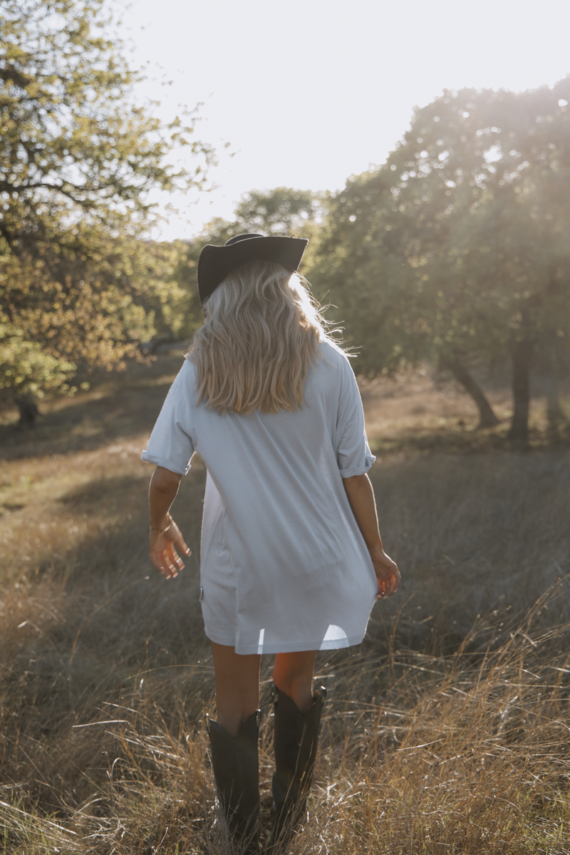 Person wearing a white dress and black boots walking through a field with trees in the background.