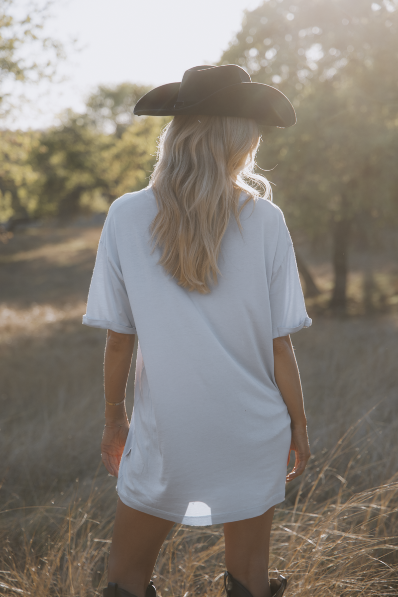 Person wearing a white t-shirt and black cowboy hat standing in a field with trees in the background.