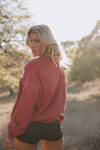 Woman in a pink sweatshirt and black shorts standing in a natural setting with trees and grass.