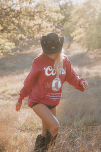 Person wearing a red sweatshirt with a logo and text, standing in a natural setting.
