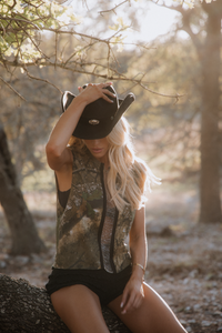 Woman sitting on a log in a forest wearing a camouflage sleeveless shirt and black shorts.