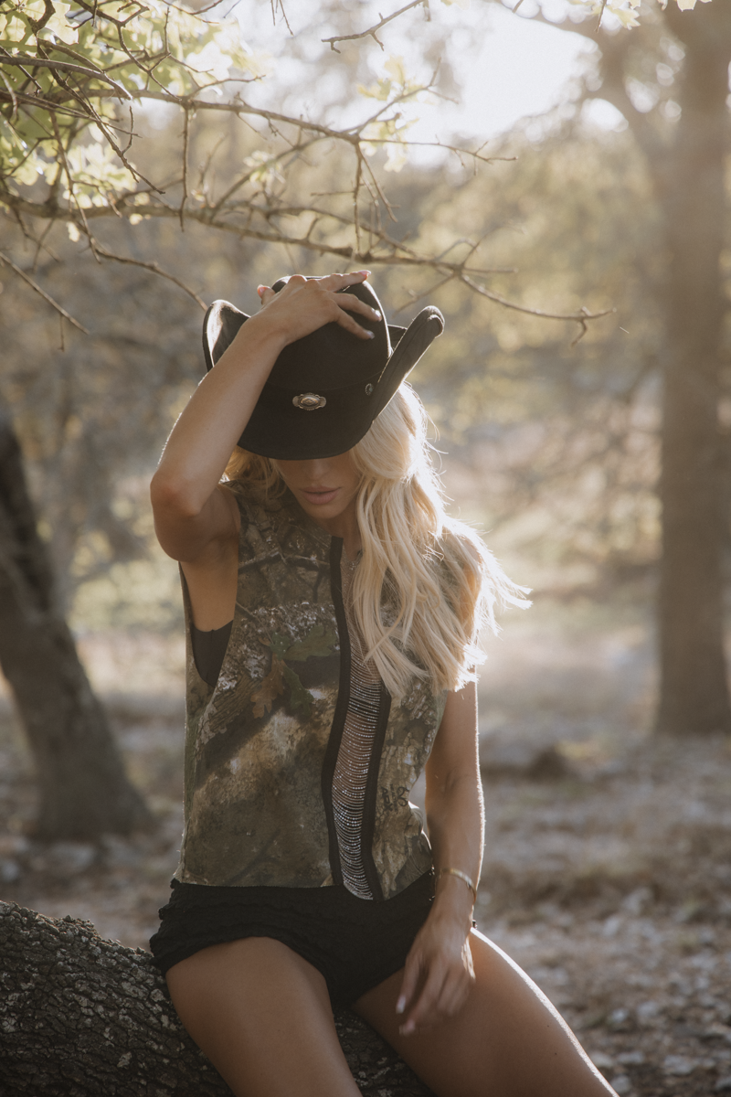 Woman sitting on a log in a forest wearing a camouflage sleeveless shirt and black shorts.