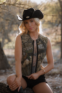Woman wearing a camouflage sleeveless jacket and black cowboy hat in a natural setting.