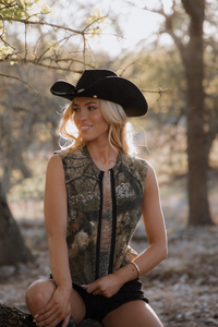 Woman wearing a black cowboy hat and camouflage vest sitting in a natural setting.