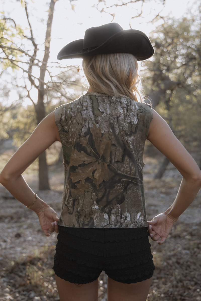 Person wearing a camouflage sleeveless shirt and black cowboy hat in a natural setting.