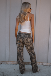 Woman wearing camouflage pants and a white top against a plain wall.